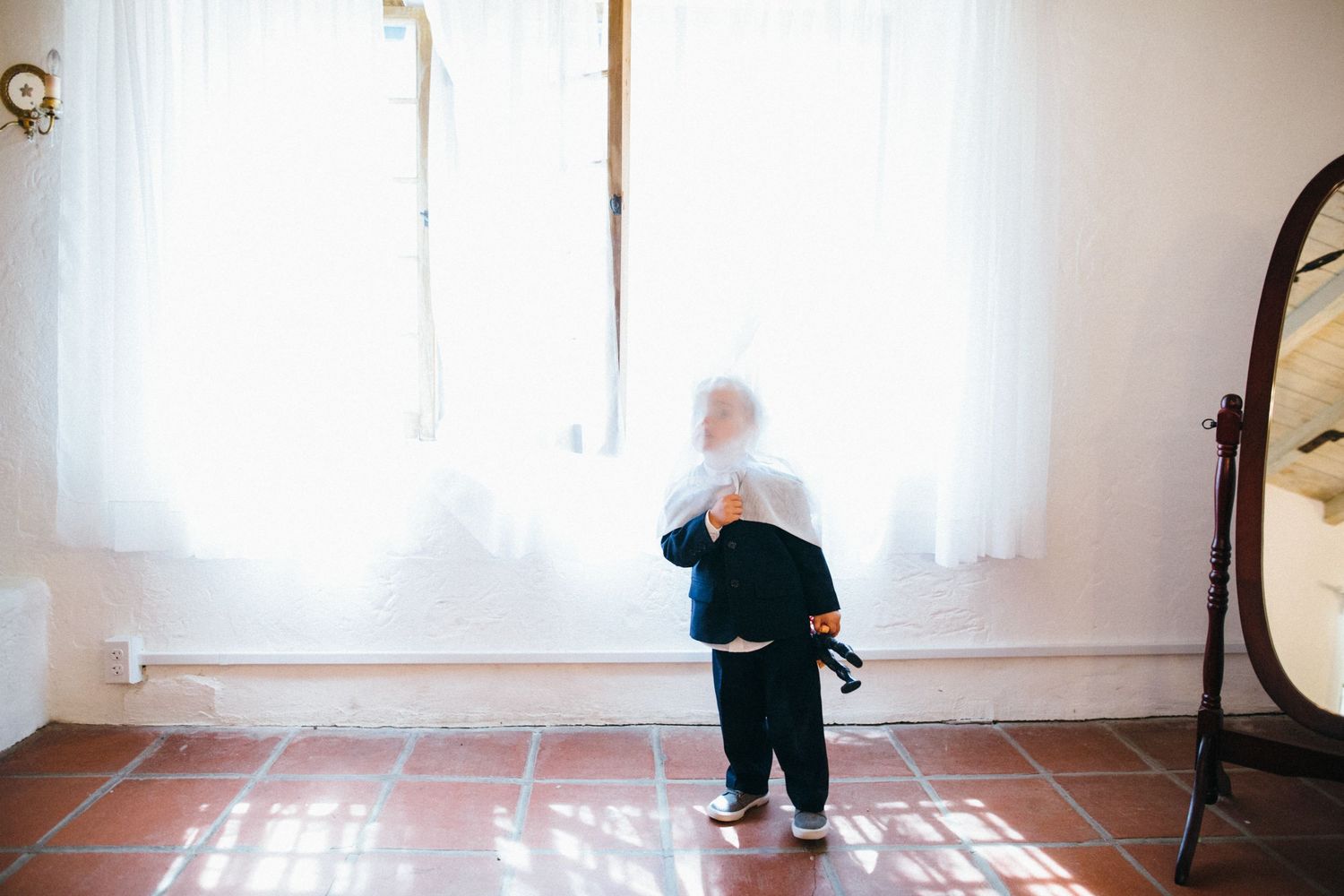 Child in tuxedo  standing by bright window with sheer curtains on tile floor near mirror at leopard carillo ranch wedding.