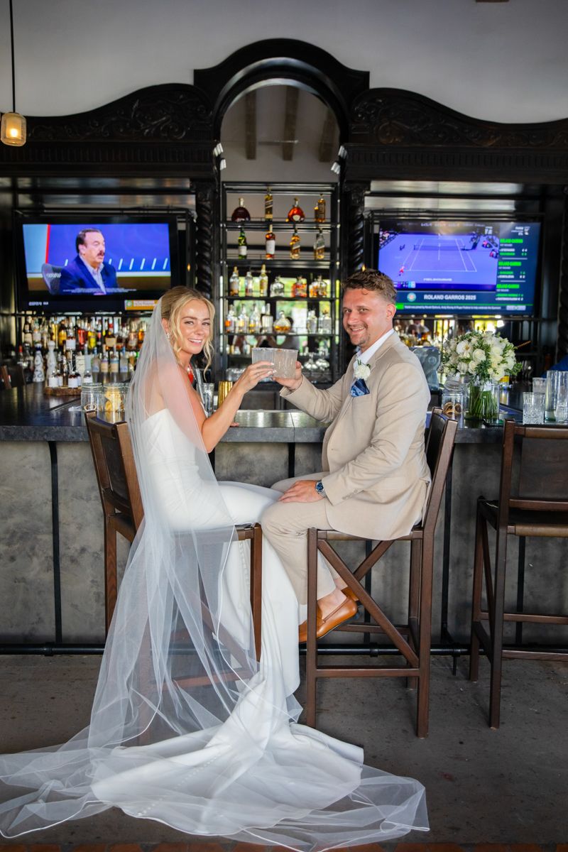 Bride and groom toasting drinks at a bar, bride wearing a long veil.