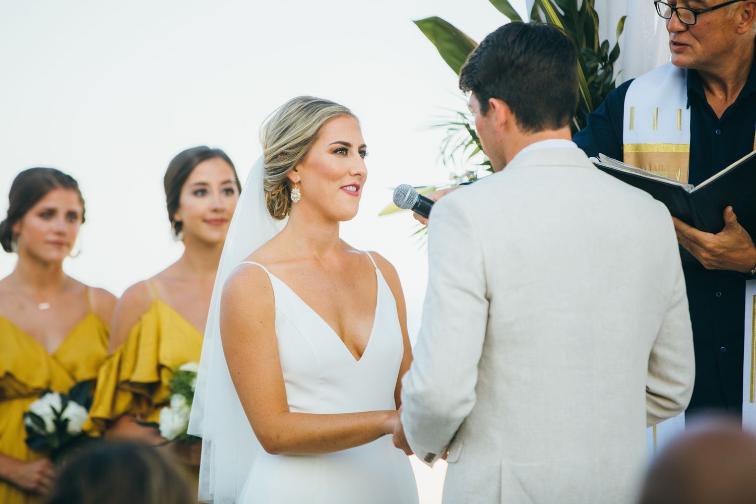 Bride in white gown exchanges vows with groom at outdoor wedding ceremony with bridesmaid in yellow dress.