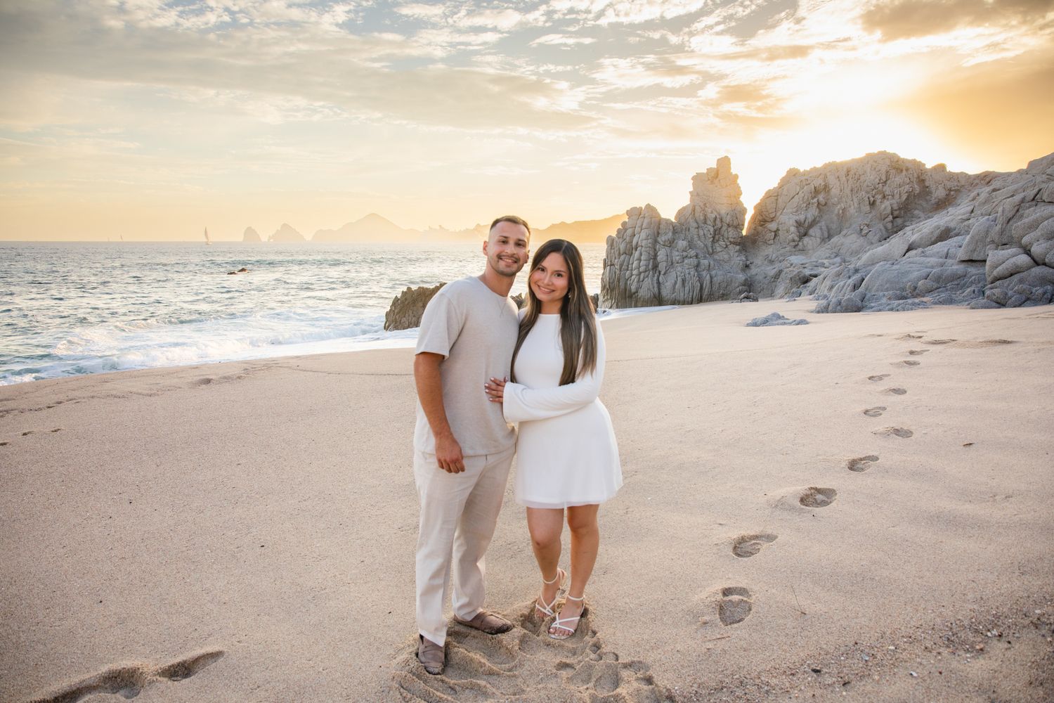 Couple in white outfits posing on sandy beach at sunset with rocky cliffs and footprints behind them.