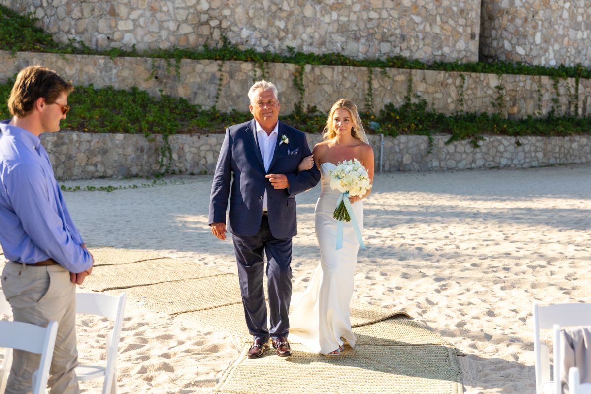 Bride walks down sandy beach aisle with father during outdoor wedding ceremony.