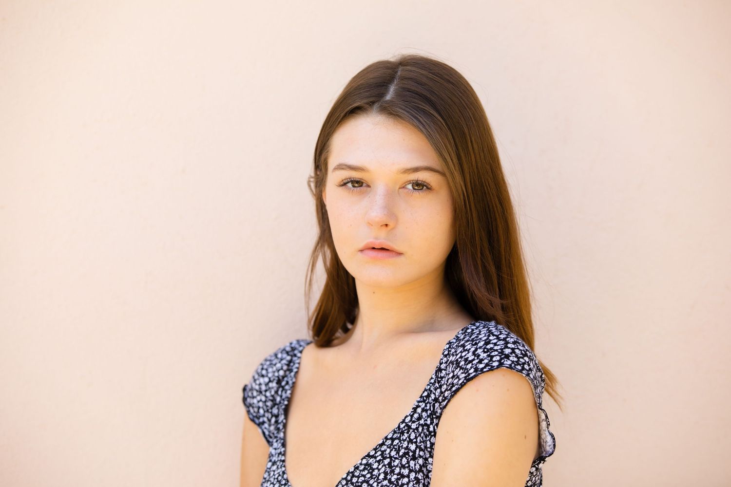 Young brunette woman in floral top posing with serious expression against a beige wall.