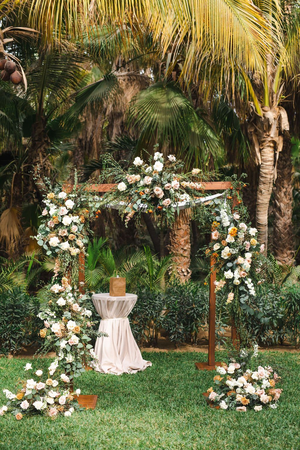 Wooden wedding ceremony arch adorned with white and peach floral arrangements in a tropical garden setting.