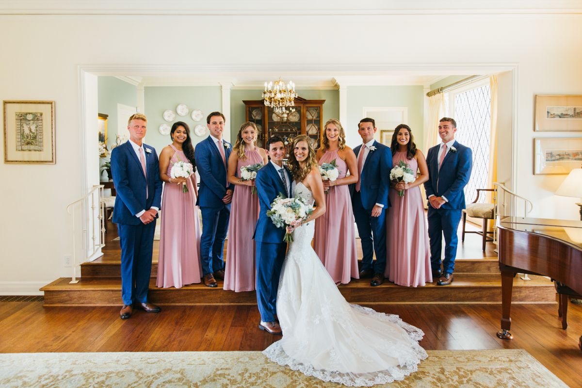 Wedding party poses indoors with bridesmaids in pink dresses and groomsmen in blue suits flanking the bride and groom.