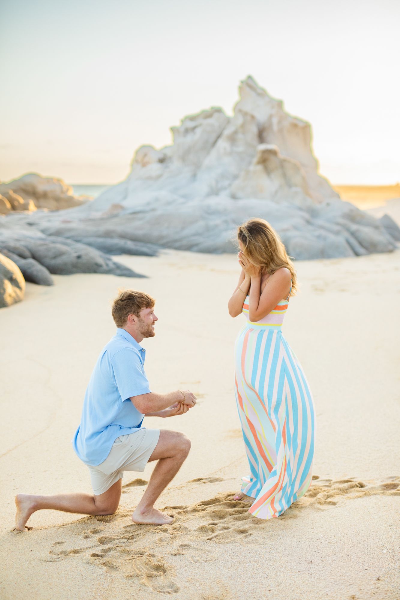 Man proposing to woman on sandy beach at sunset with white rock formation backdrop, woman covers face in surprise in cabo