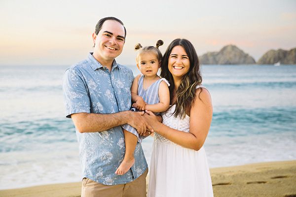Young couple holding toddler on a beach with ocean and rocky cliffs at sunset.