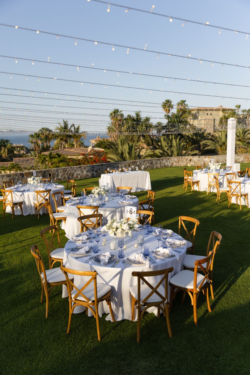 Outdoor wedding reception on green lawn with round tables, string lights, and ocean view.