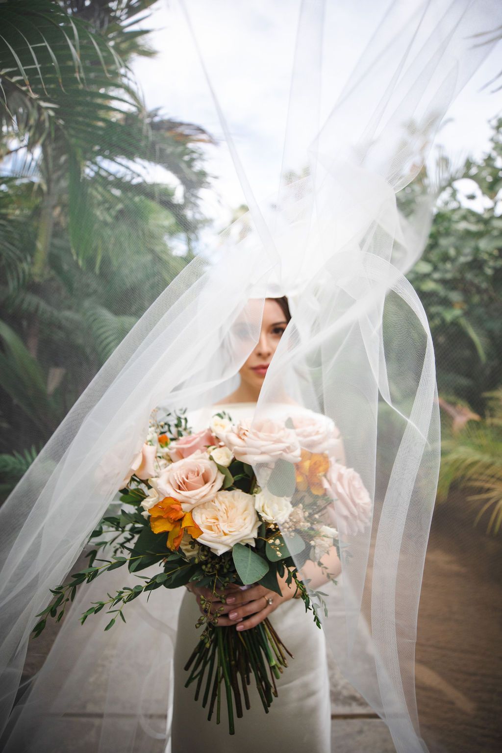 Bride with flowing veil holding colorful bouquet of pink, peach and orange roses outdoors.