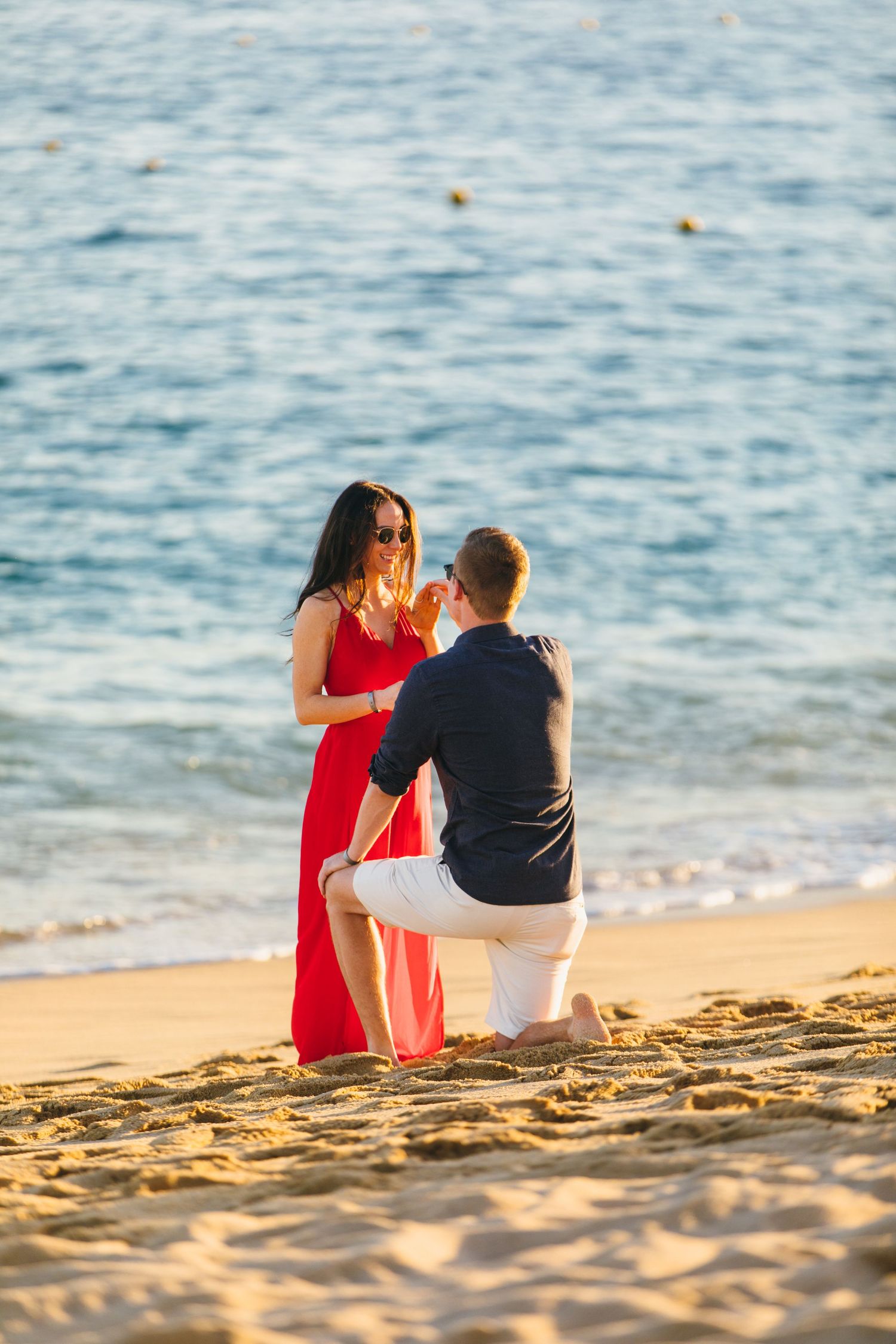 Man proposing on one knee to a woman in a red dress on a sandy beach at sunset.