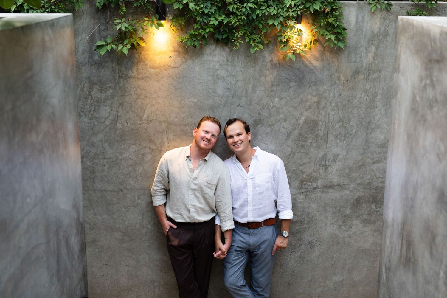 Two smiling men pose together against a concrete wall with green ivy and warm lighting overhead.