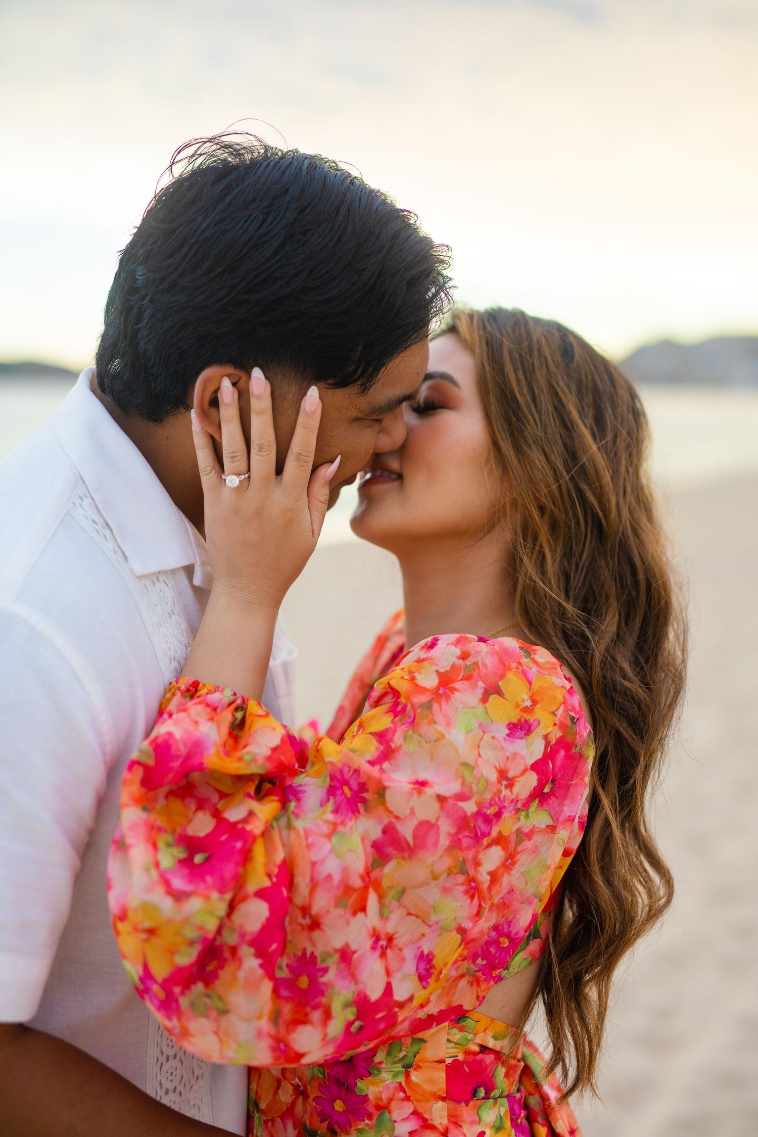 Couple sharing a romantic kiss on the beach, woman in colorful floral dress showing engagement ring.