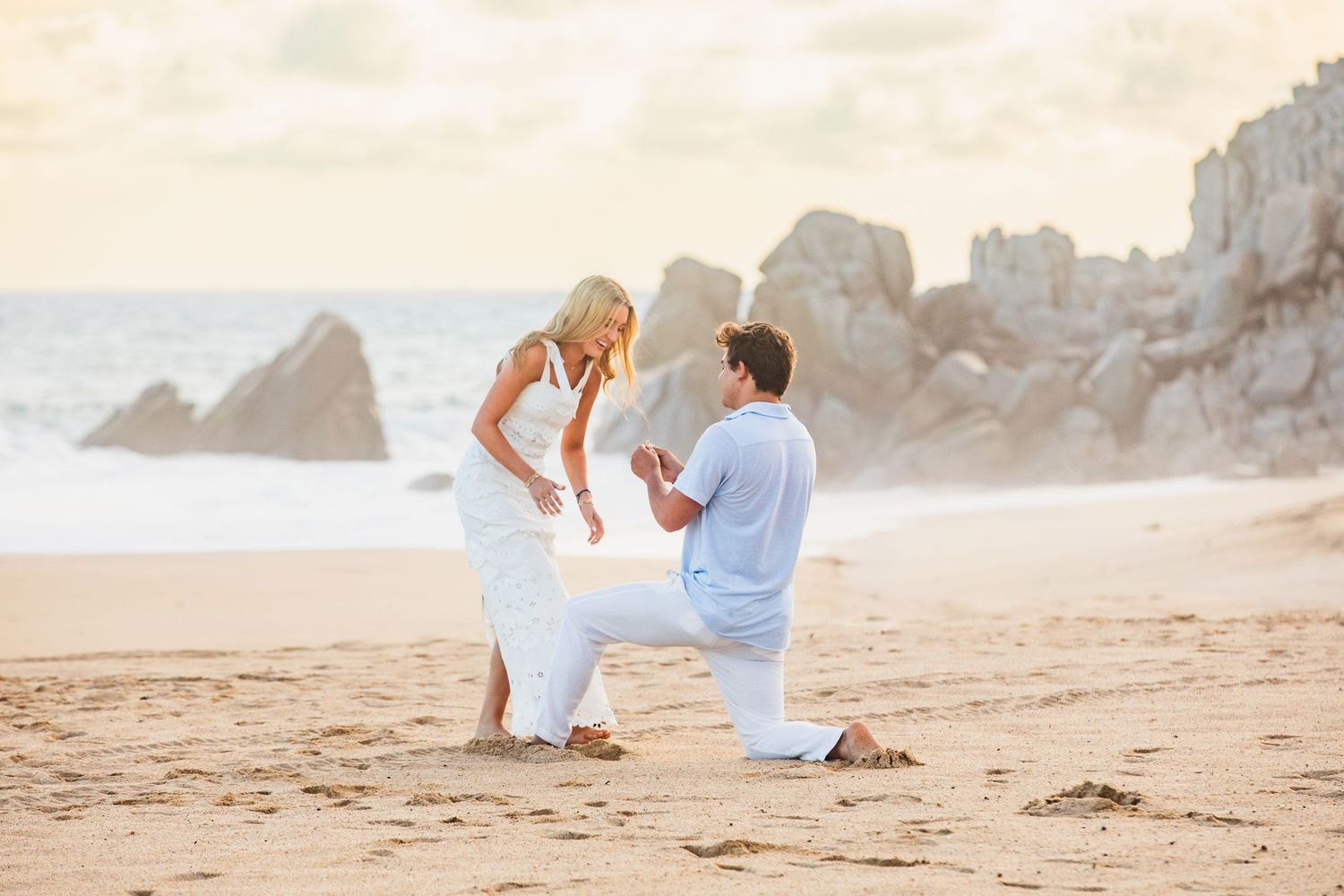 Man proposing on one knee to woman in white dress on a beautiful sandy beach at sunset.
