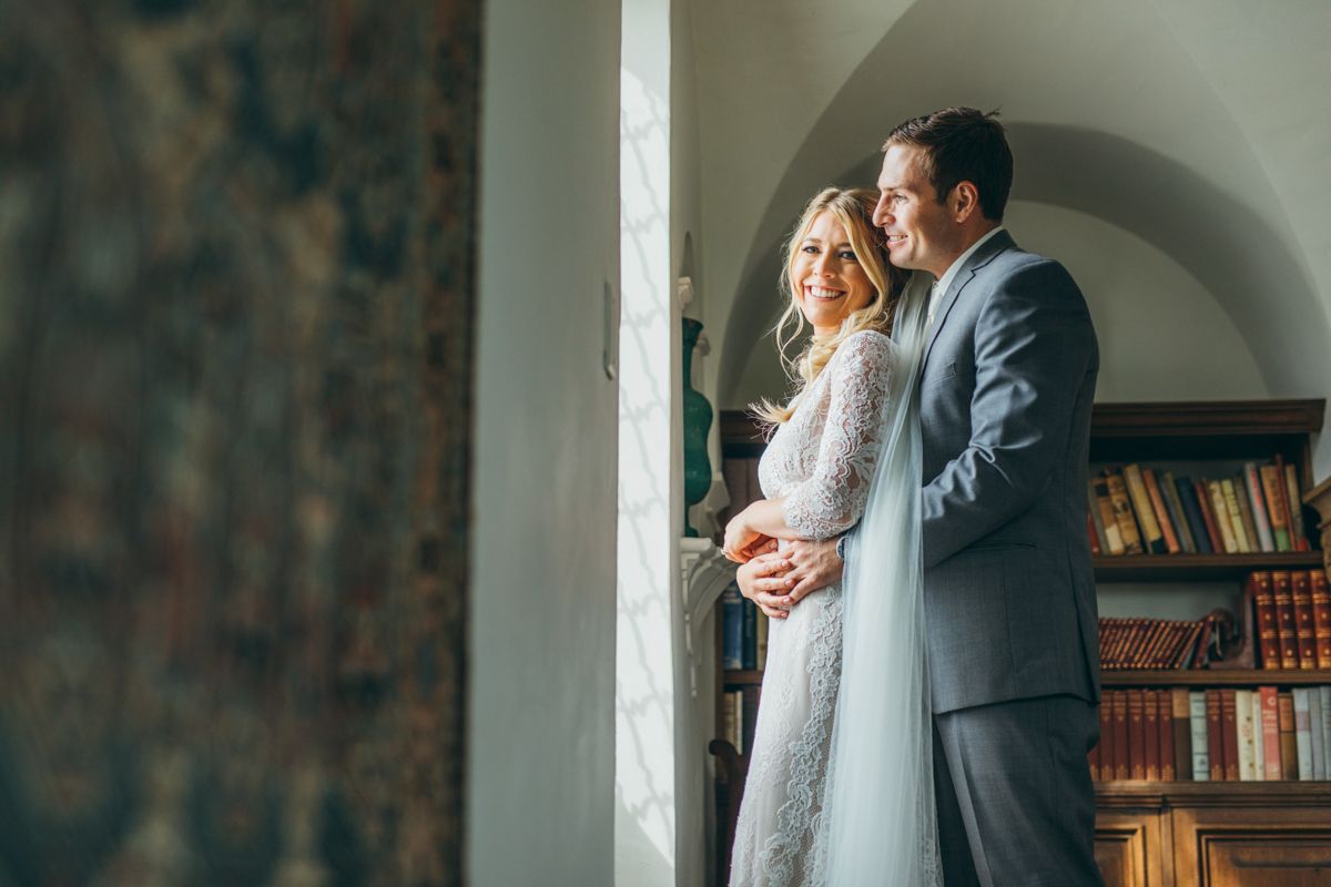 Bride and groom share intimate moment by window in elegant library with bookshelves.