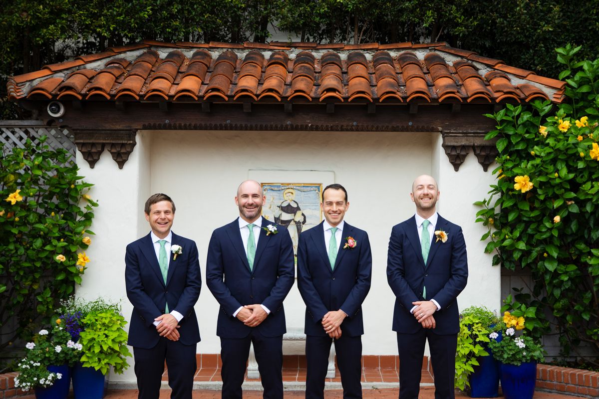 Groom and three groomsmen in navy suits with mint ties pose before Spanish-style terracotta roof.
