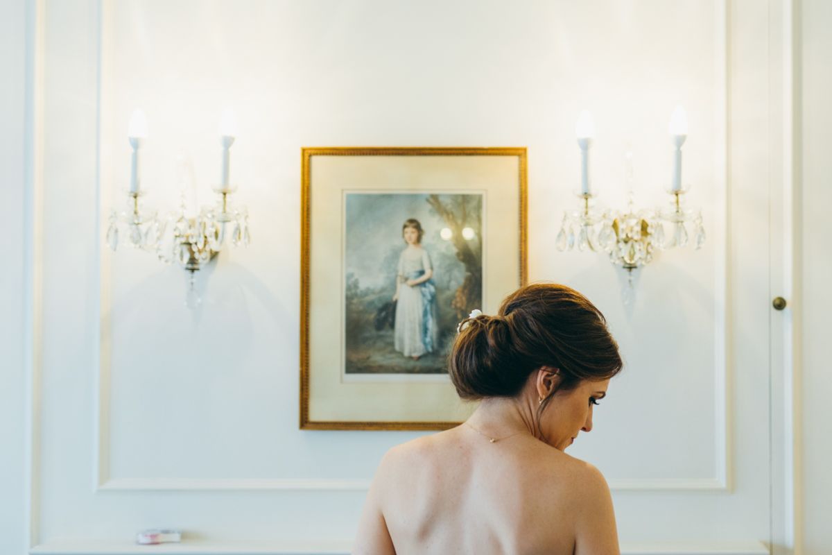 Bride with elegant updo standing before a framed portrait flanked by crystal wall sconces in a classic interior.