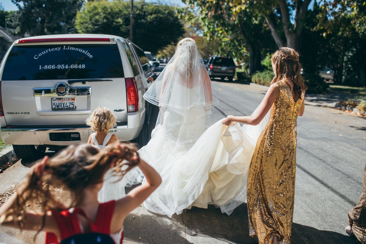Bride in white gown and veil walking outdoors, assisted by bridesmaid in gold dress and flower girl.