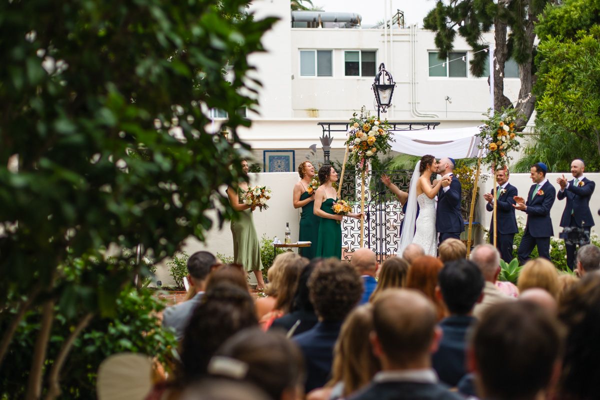 Couple kisses under floral chuppah during outdoor wedding ceremony as guests watch from seats.