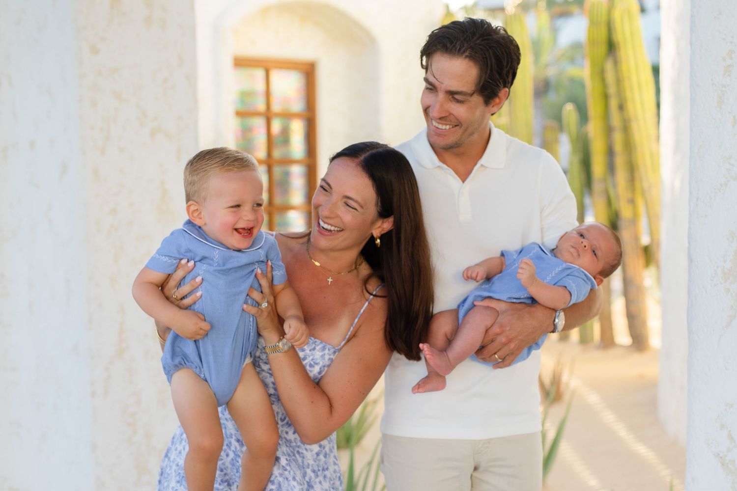 Happy family with two babies posing outdoors, smiling couple holding infant twins in blue outfits near white columns.