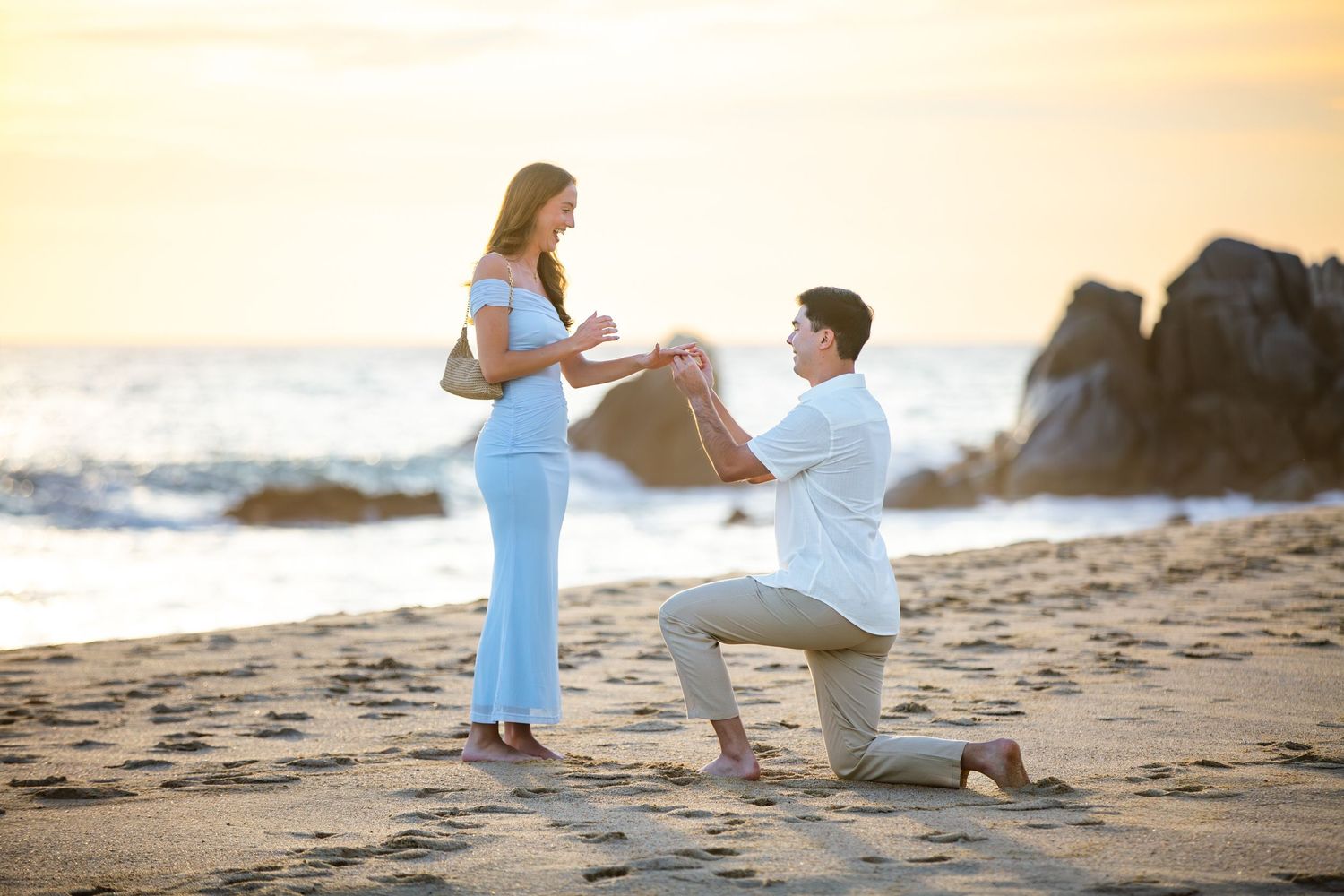 Man proposing to woman on a sandy beach at sunset with ocean waves and rocky cliffs in the background.