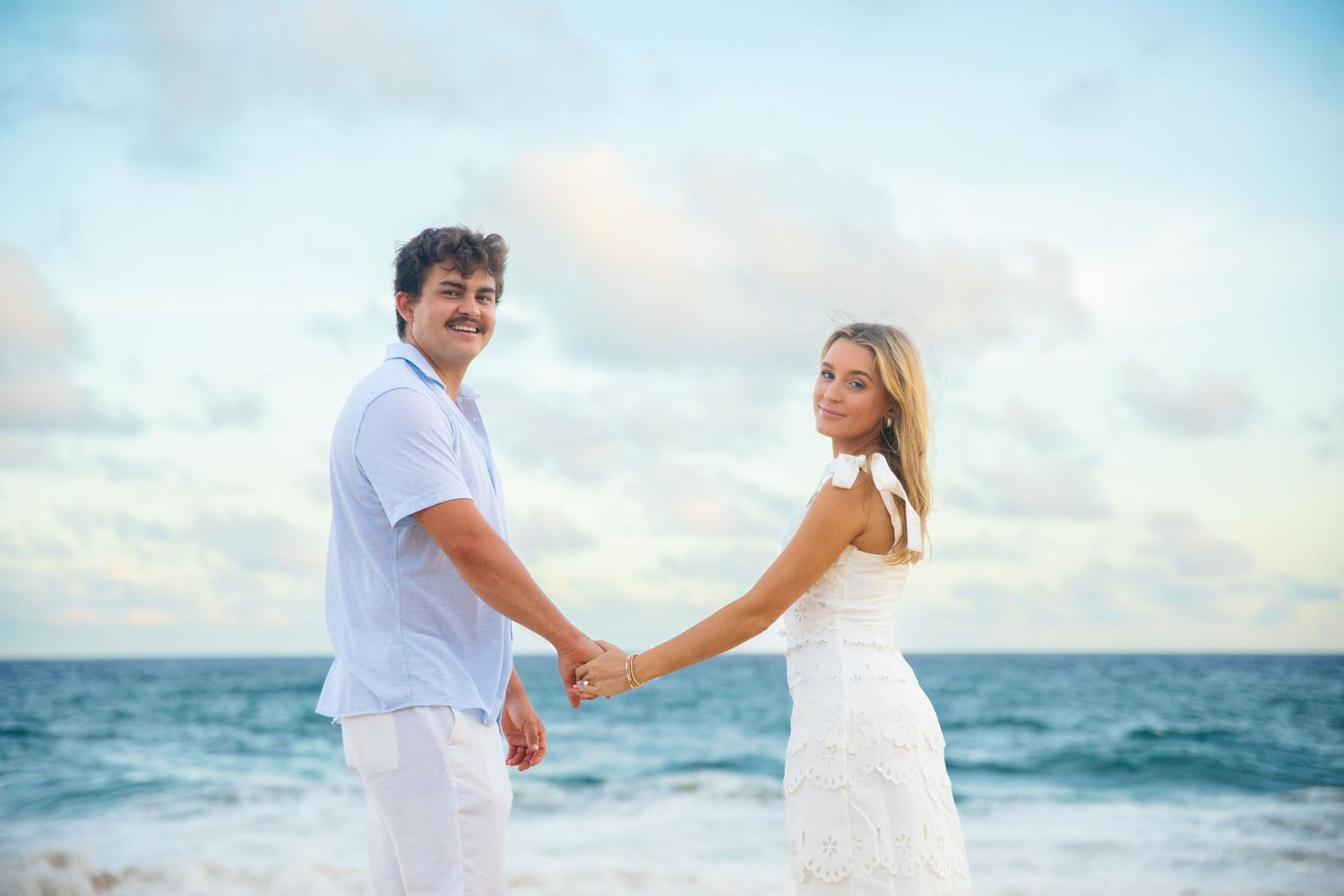 Couple holding hands on a tropical beach at sunset, woman in white dress, man in white shirt.