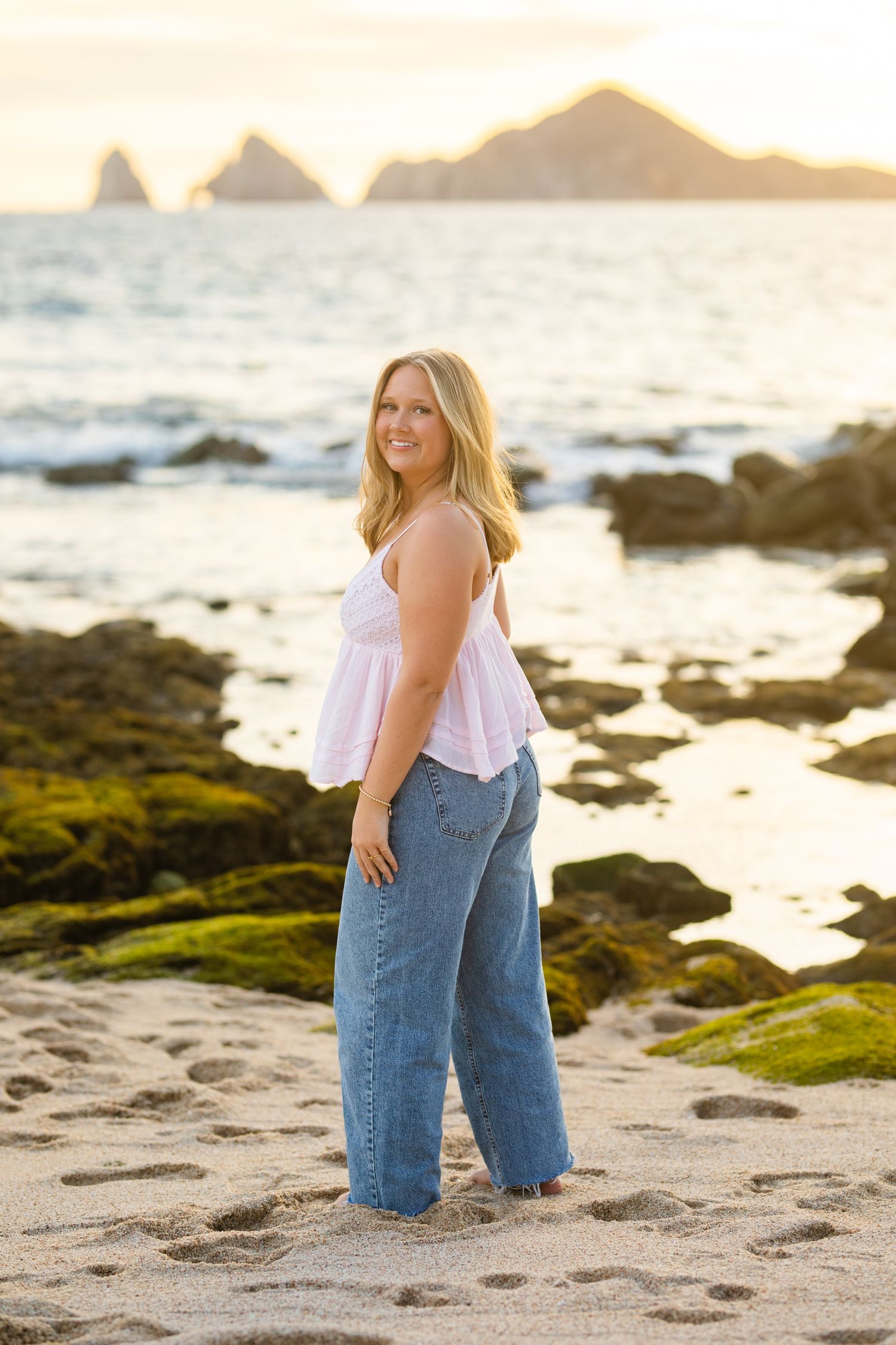 Smiling blonde woman in pink top and jeans posing on a rocky beach at golden sunset.
