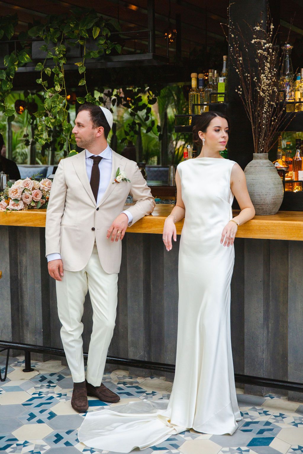 Stylish bride and groom posing at a tropical bar, groom in beige suit and bride in sleek white gown.