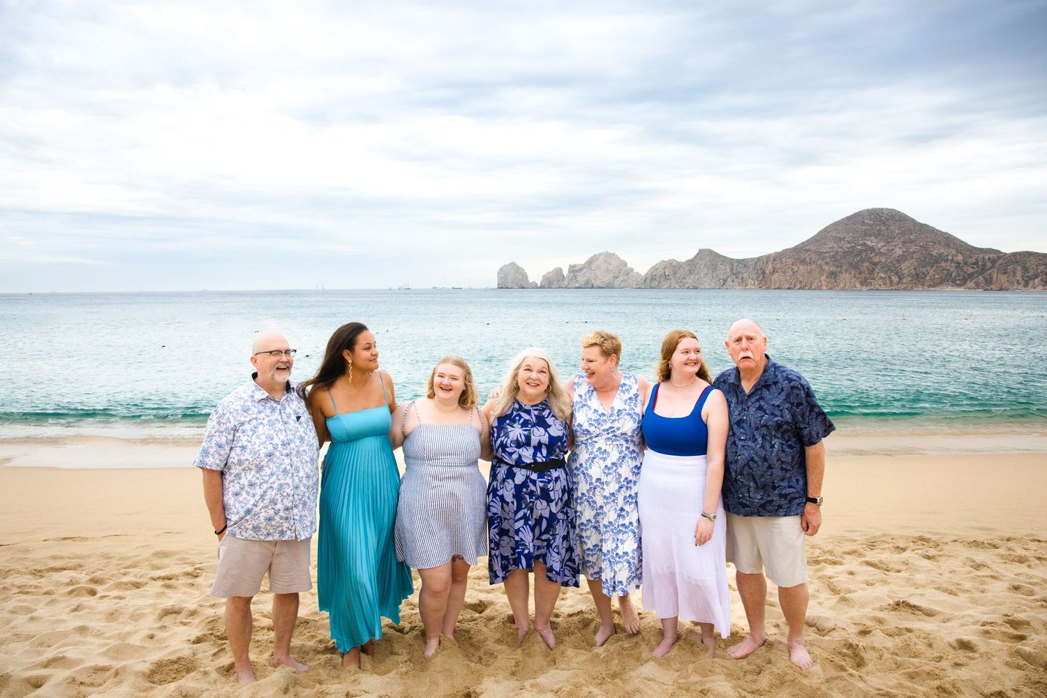 Group of seven adults posing together on a sandy beach in Cabo San Lucas with turquoise water and rocky cliffs in the background.