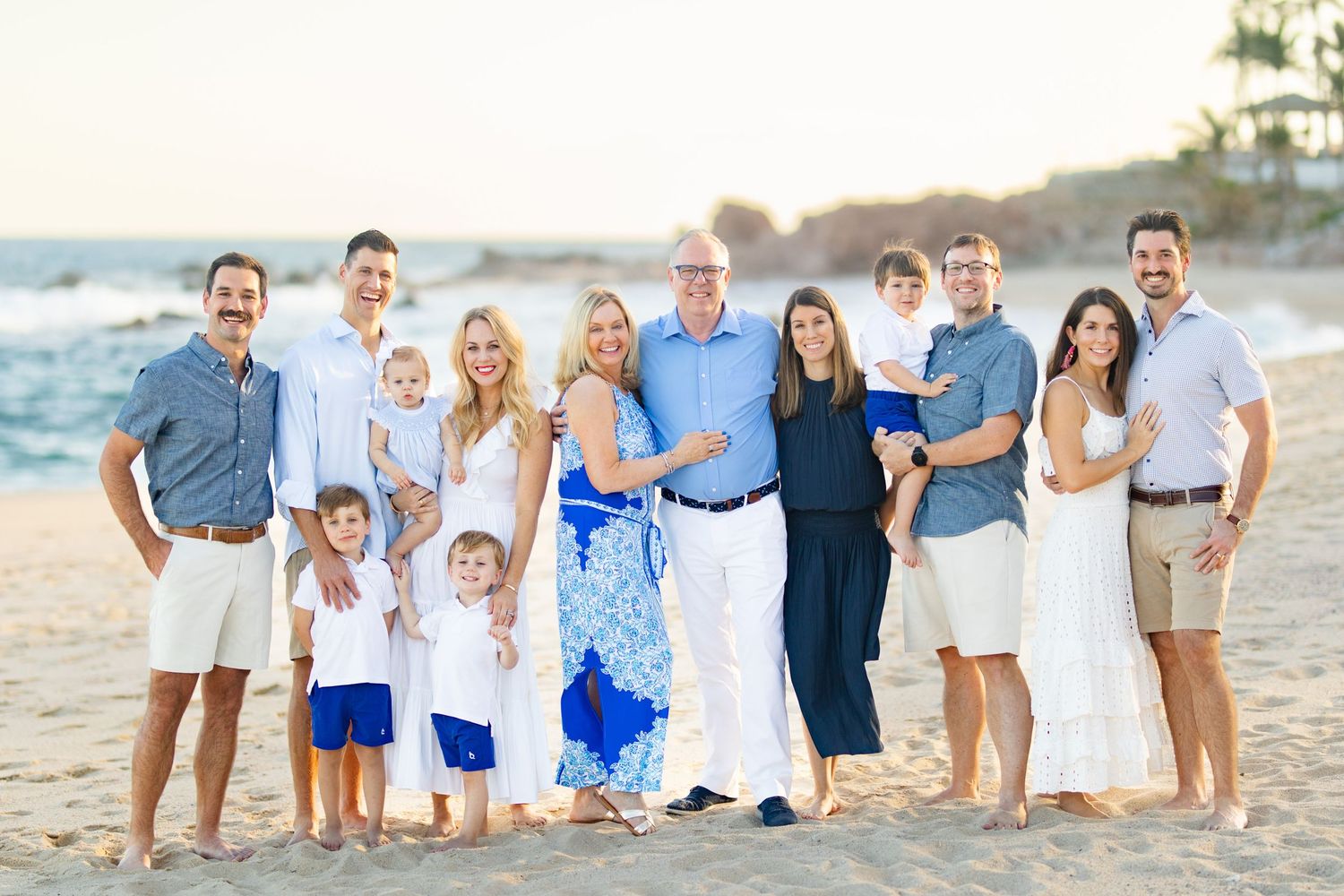 Large family group portrait on a sandy beach at sunset, dressed in coordinating white and blue outfits.