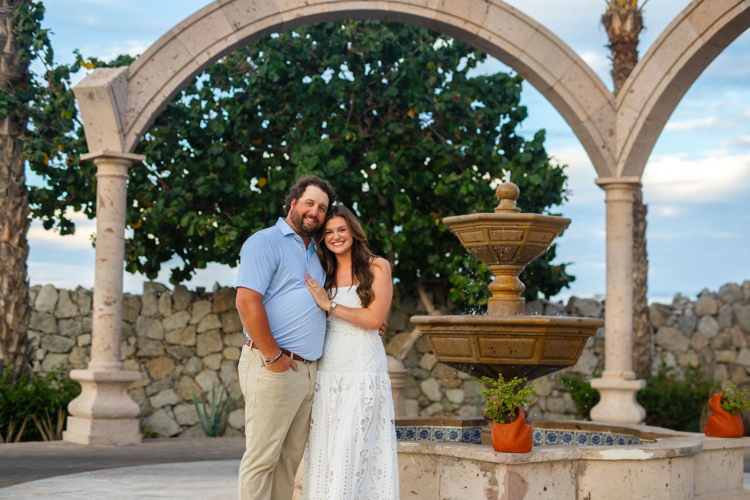Couple smiling together under stone arches with a fountain and tropical trees at an outdoor venue.