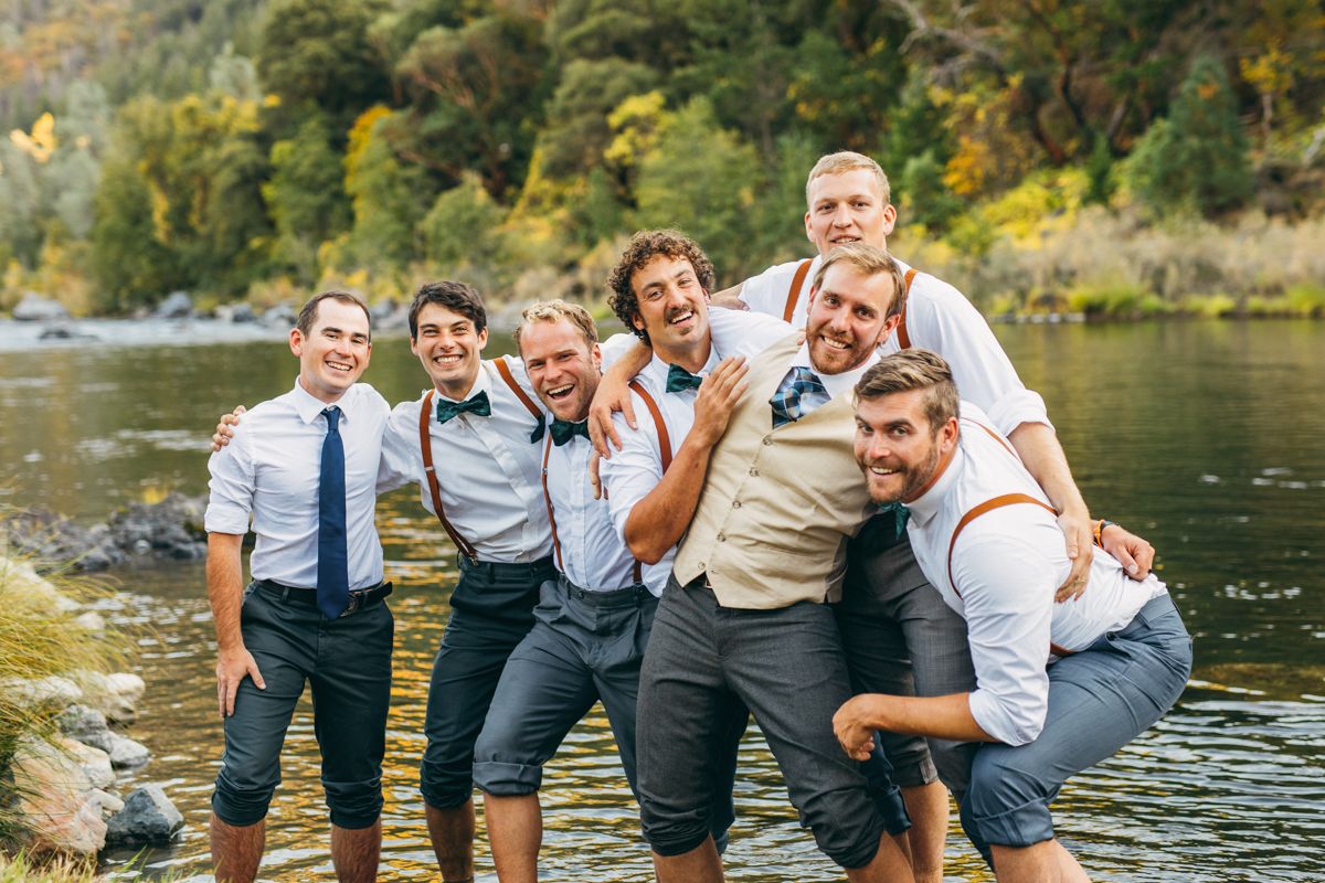 Groom and groomsmen in white shirts and suspenders posing playfully by a river outdoors.