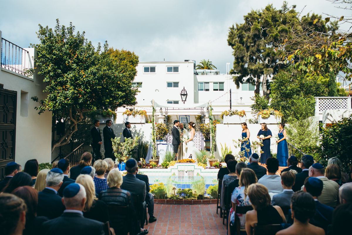 Outdoor Jewish wedding ceremony with guests seated in courtyard of a white Spanish-style building.