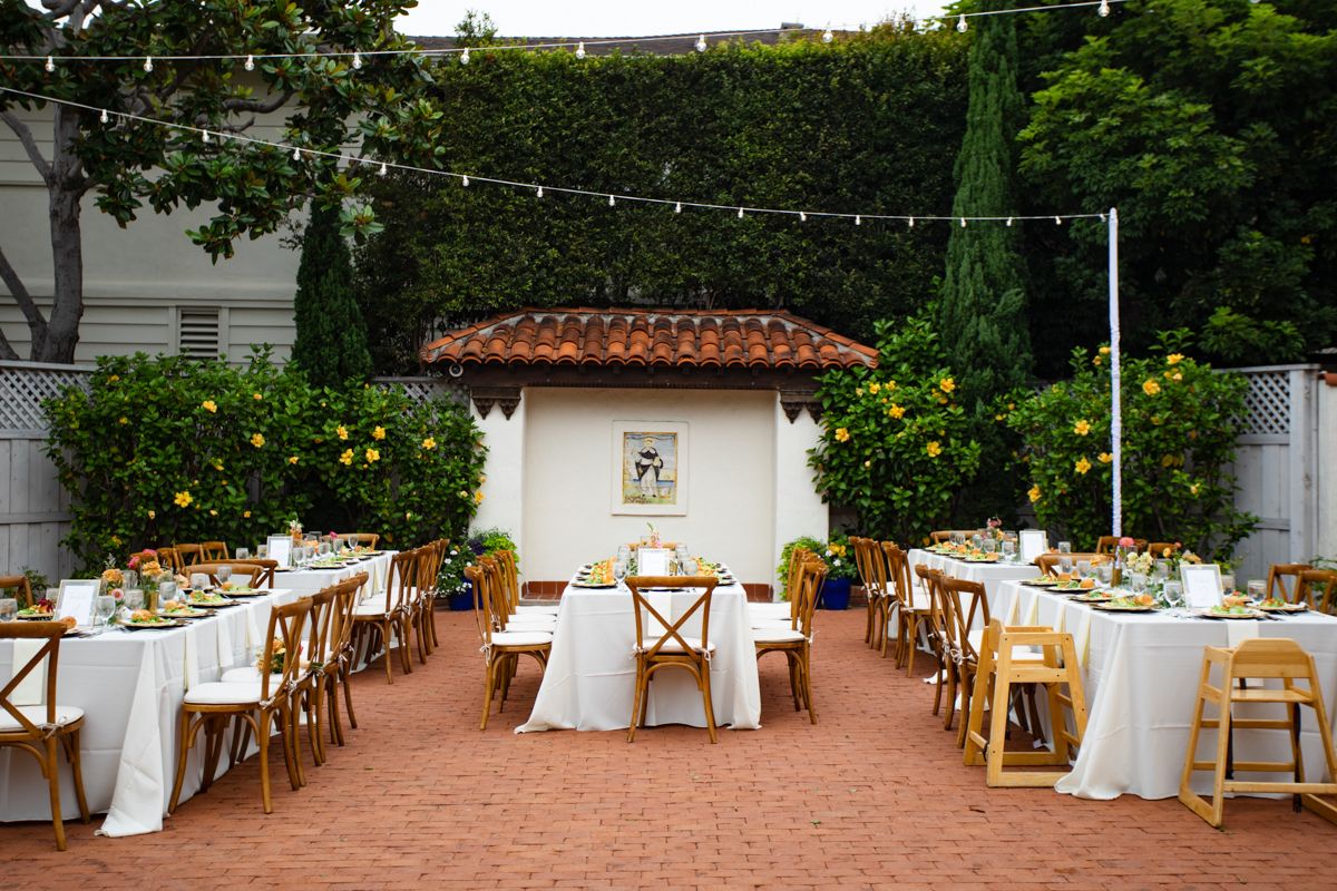 Elegant outdoor wedding reception with long white-draped tables, wooden chairs, string lights, and lemon trees in a courtyard.