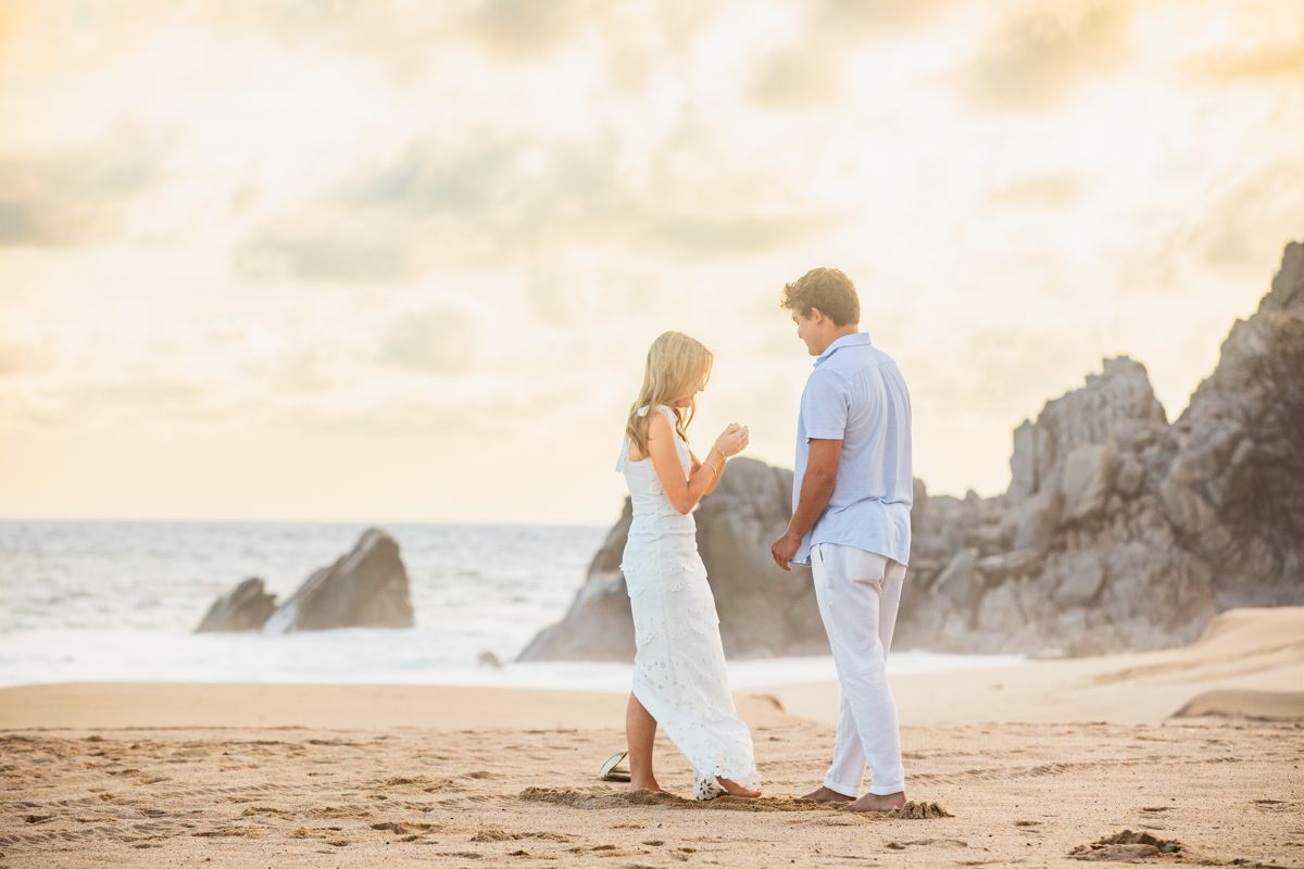 Couple holding hands and facing each other on a scenic beach at sunset with rocky cliffs.