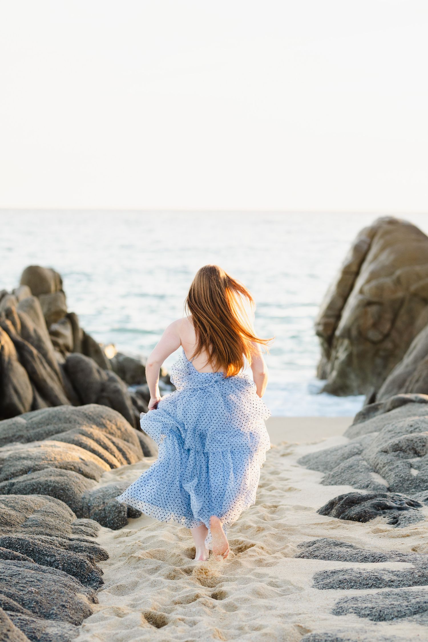 Redhead woman in blue floral dress walking barefoot toward ocean between rocky beach boulders.