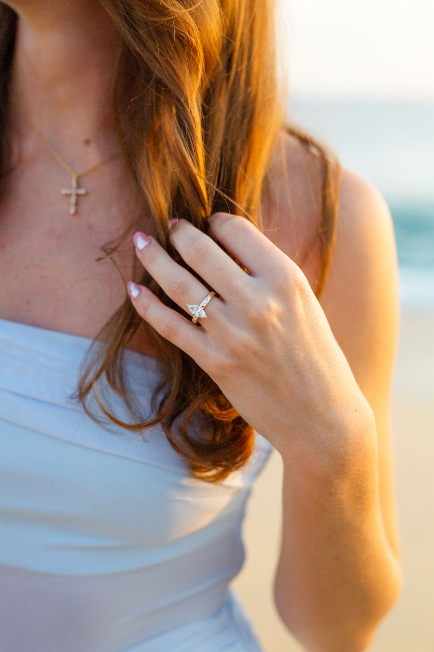 Woman in white dress displaying diamond engagement ring, wearing cross necklace, with auburn hair at beach.