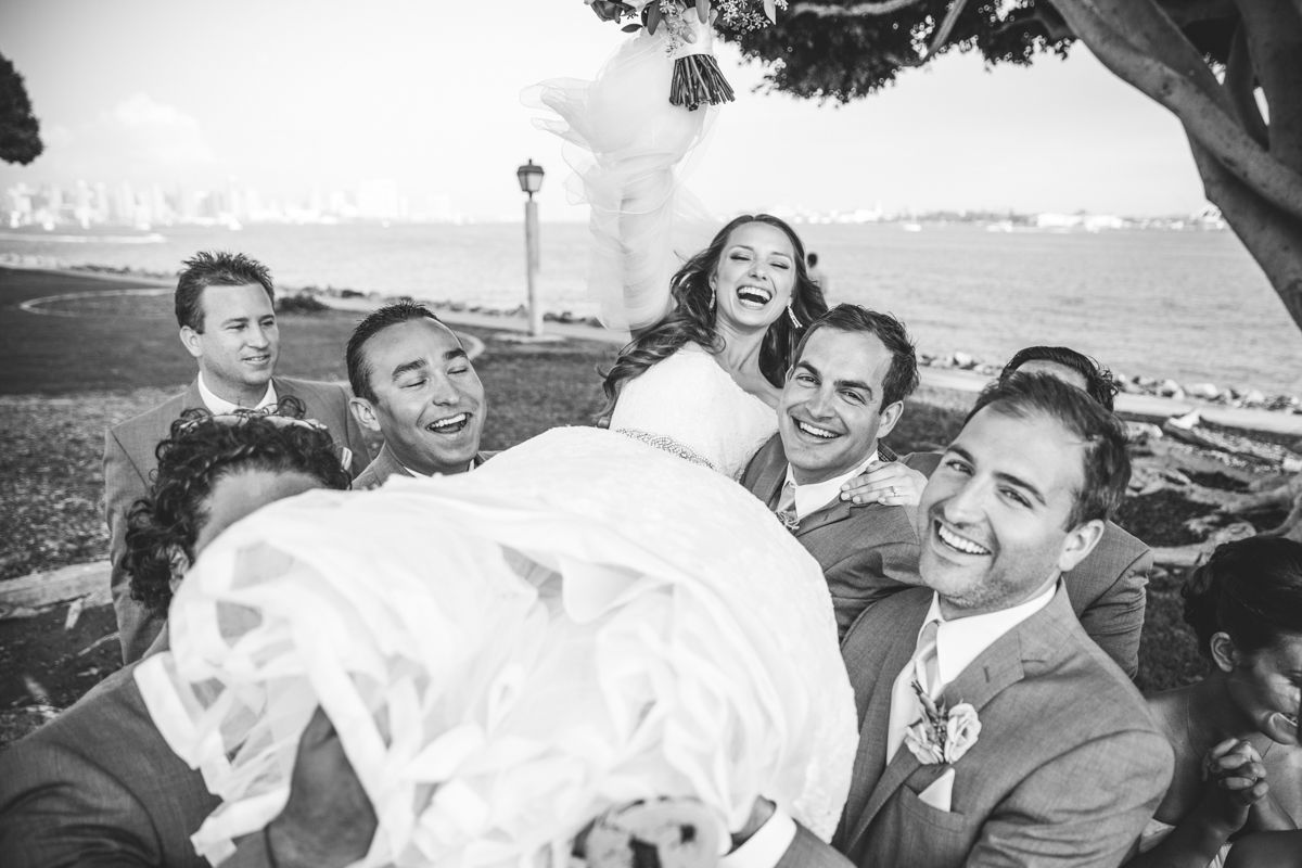 Black and white photo of laughing bride being lifted by groomsmen in suits near waterfront.