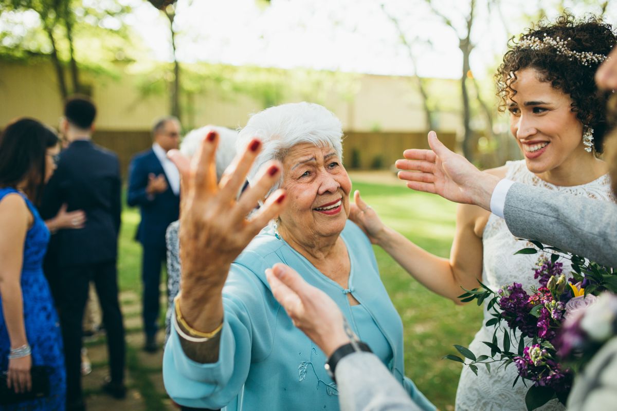 Elderly woman raising hand joyfully as bride with bouquet greets her at outdoor wedding.