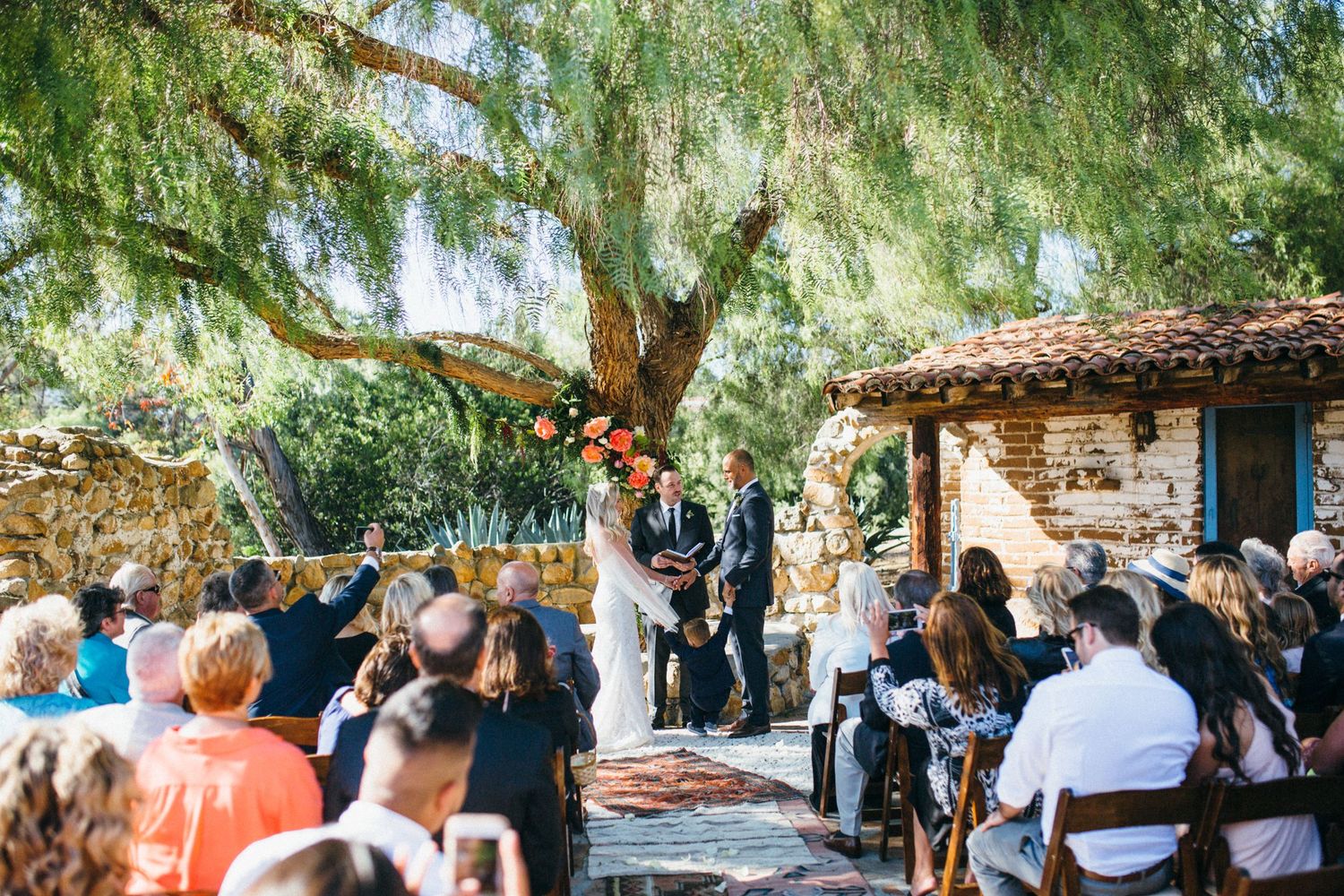 Outdoor wedding ceremony under a large tree with guests seated, officiant, and couple at a rustic Leo Carrillo ranch