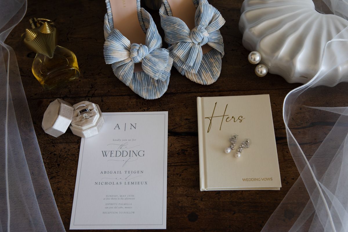 Wedding flat lay with blue heels, invitation, veil, rings, and pearl accessories.