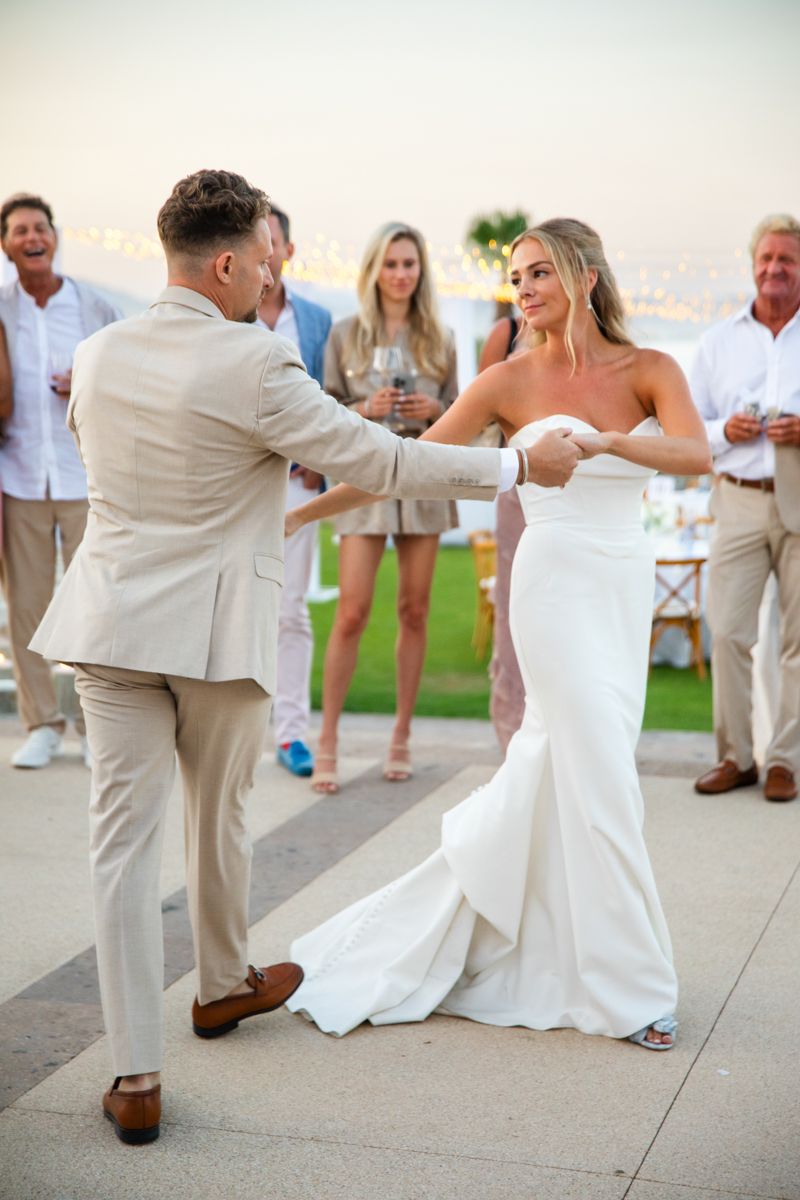 Groom twirling bride in white fitted gown during outdoor wedding reception at dusk.