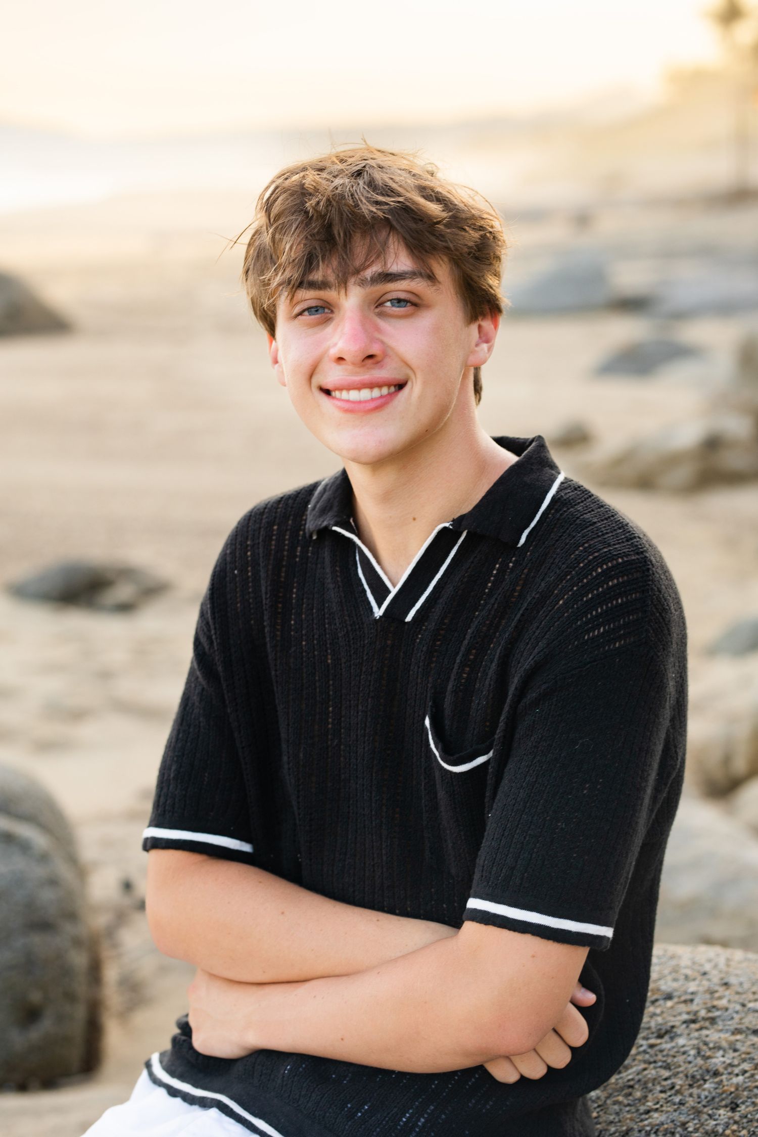 Smiling teen boy in black knit polo sitting cross-armed on beach rocks at golden hour.