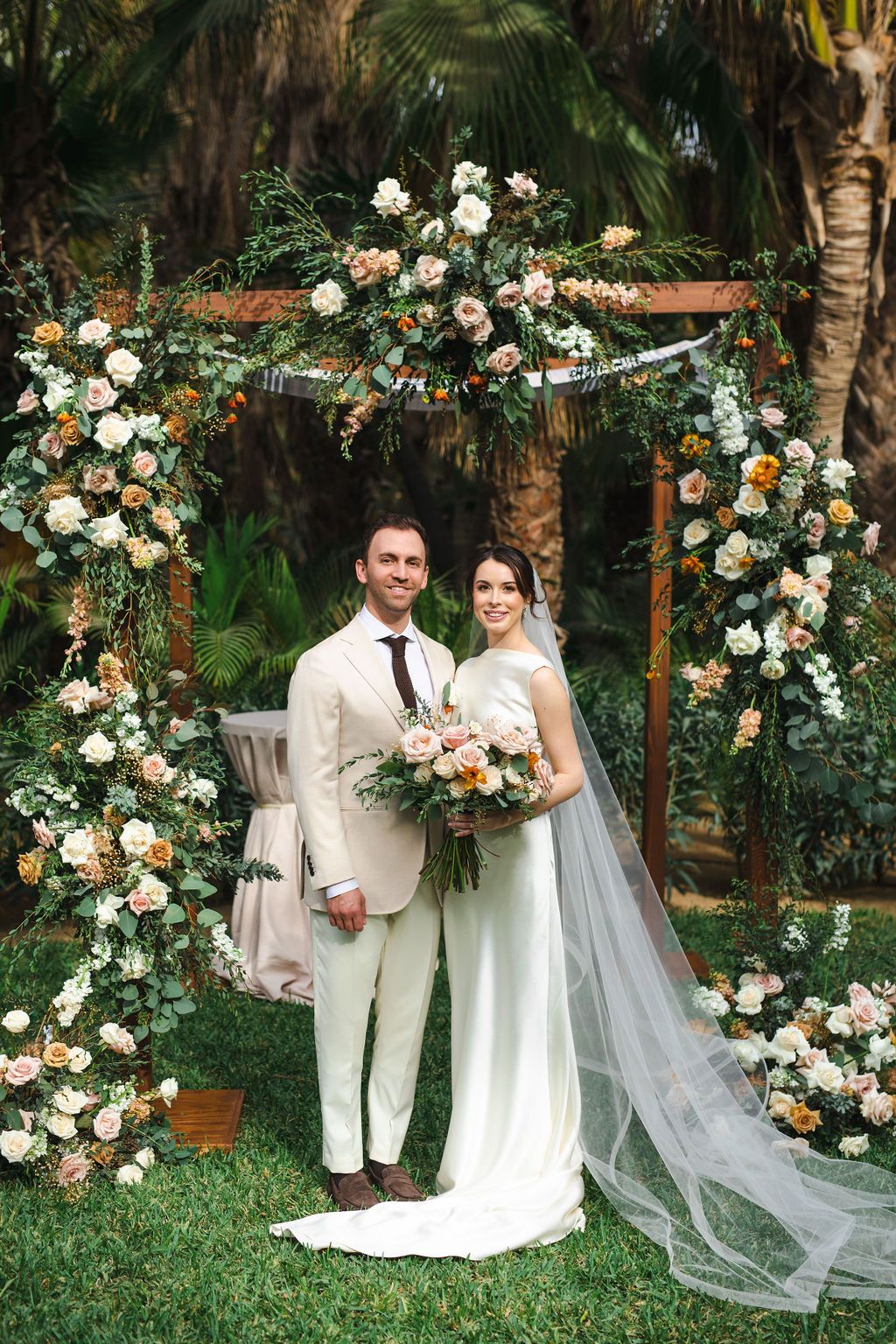 Bride and groom posing under a floral arch with roses and greenery at an outdoor garden wedding ceremony.