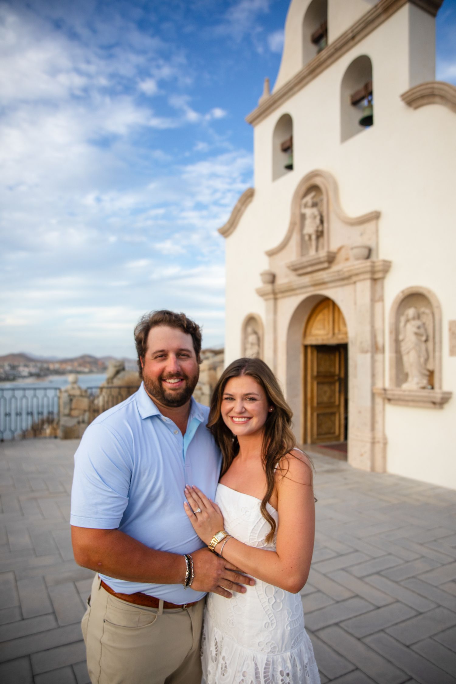 Smiling couple poses in front of a white Spanish colonial church in Cabo San Lucas on a sunny day.
