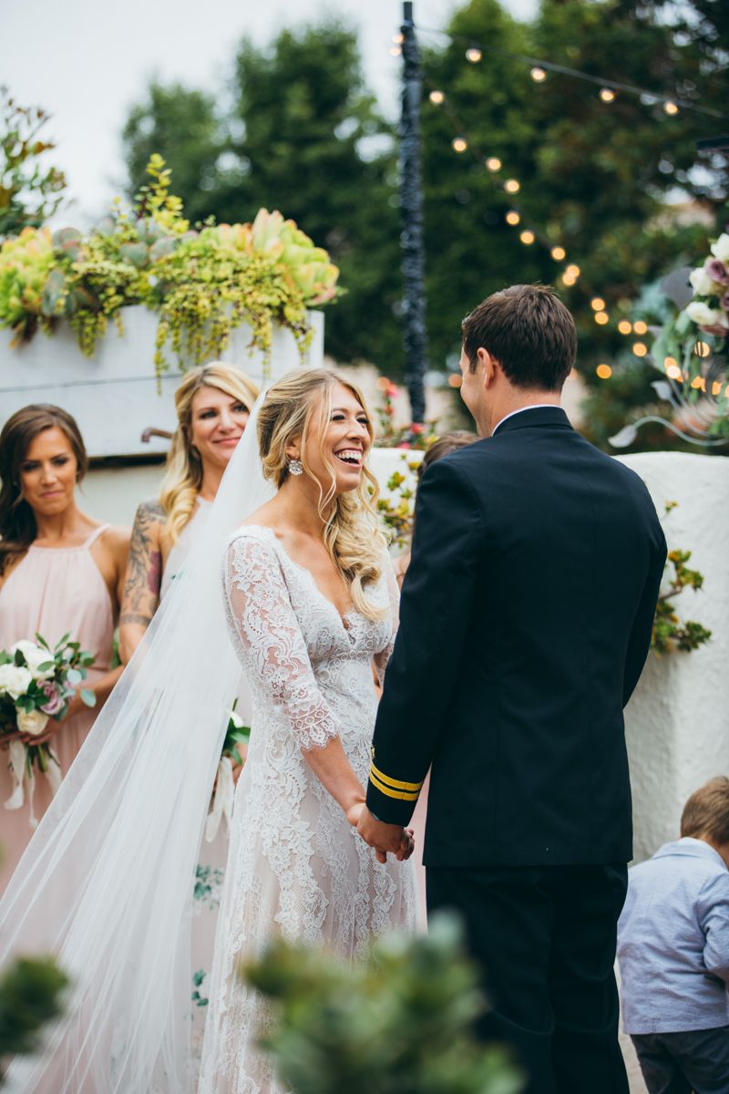 Laughing bride in lace gown holds grooms hands during outdoor wedding ceremony with bridesmaids.