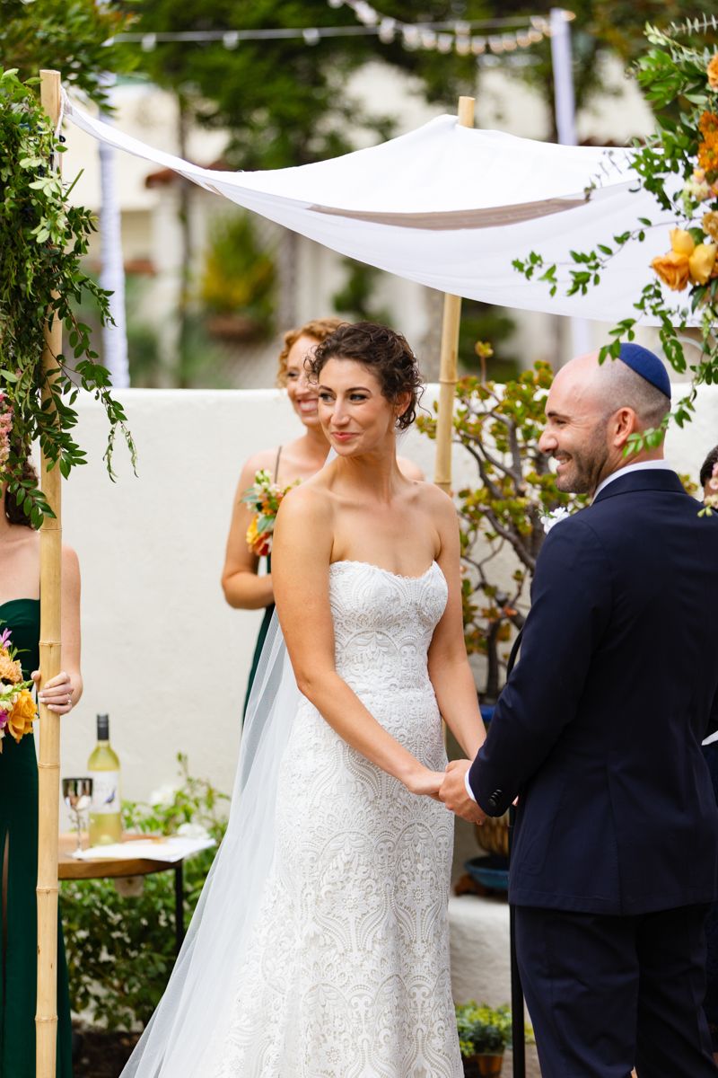 Bride smiles at groom during outdoor Jewish wedding ceremony under white chuppah with floral decor.