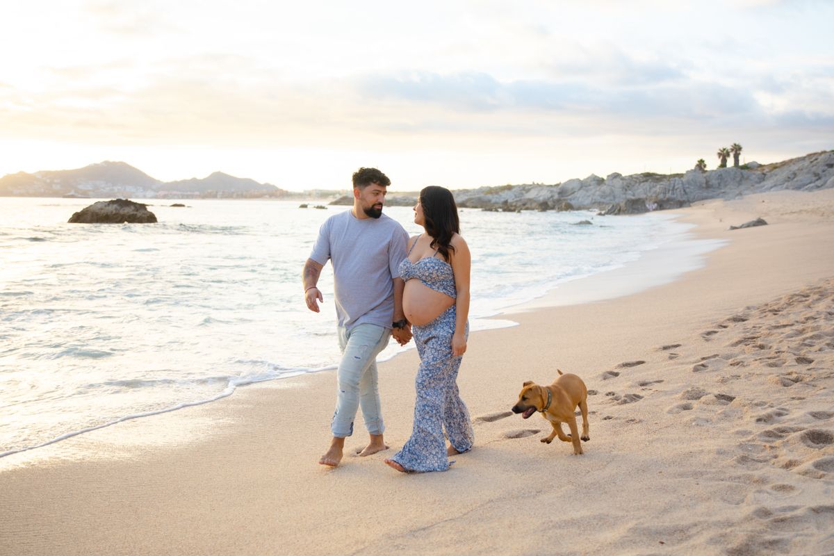 Expecting couple walks along sandy beach at sunset with their dog, woman cradling pregnant belly.