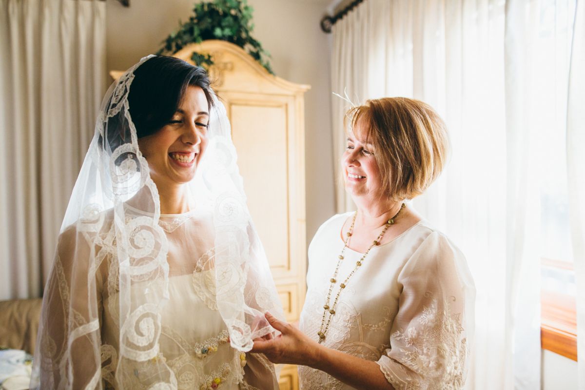 Mother helps bride in lace wedding dress and veil prepare for ceremony in sunlit room.