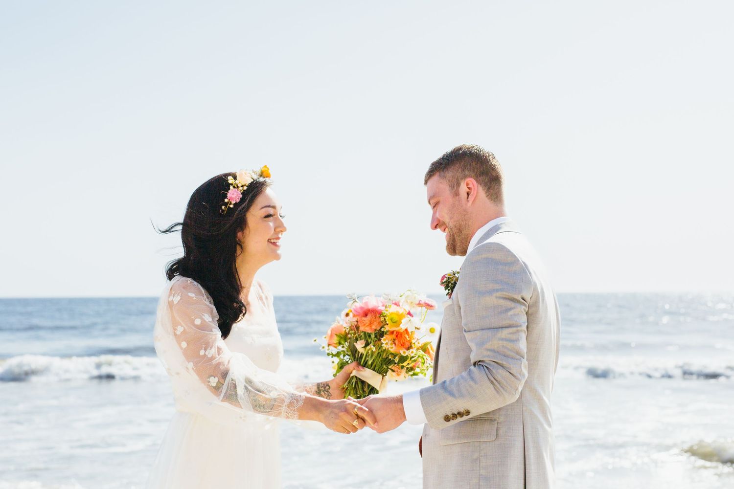 Bride and groom facing each other holding a bright bouquet on a sunny beach.