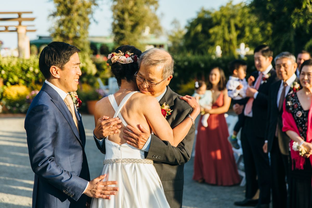 Emotional bride embraces family member while groom stands nearby during outdoor wedding ceremony.