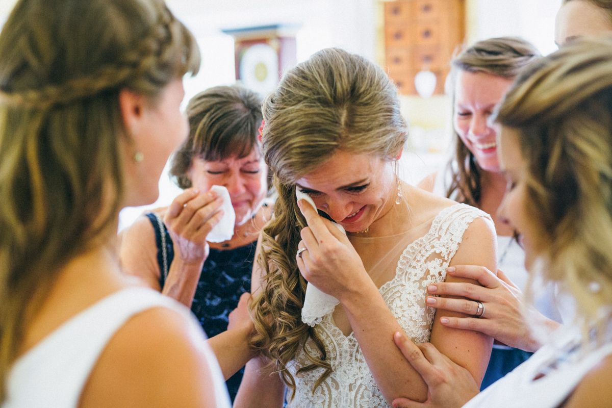 Emotional bride wiping tears surrounded by supportive bridesmaids and mother before ceremony.